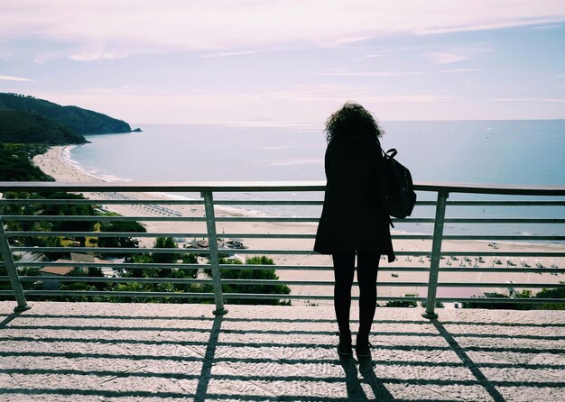 Foto vista posteriore di una donna a silhouette in piedi accanto a una ringhiera contro il mare