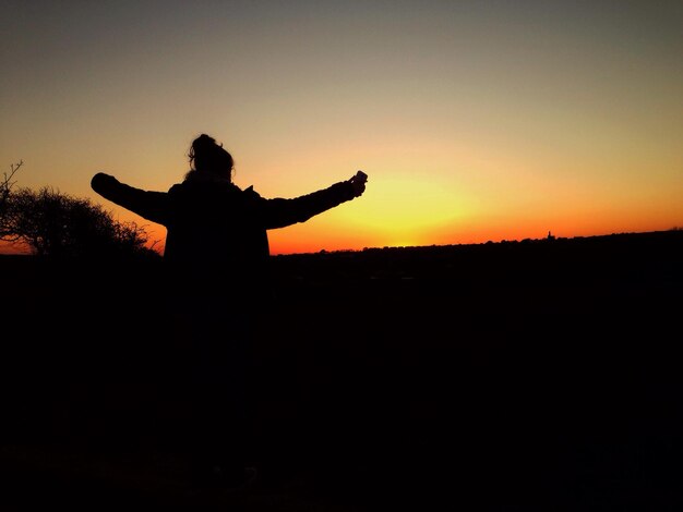 Rear view of silhouette woman with arms outstretched standing on field at sunset