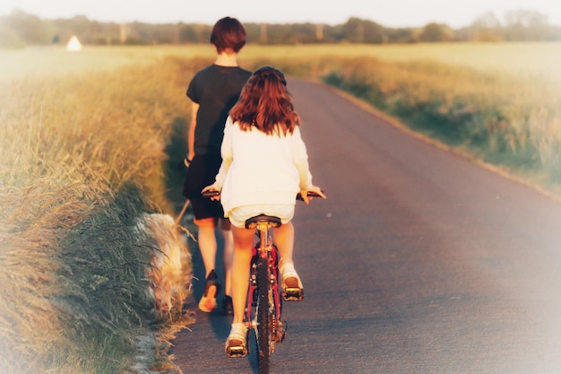 Photo rear view of sister riding cycle by brother walking on road