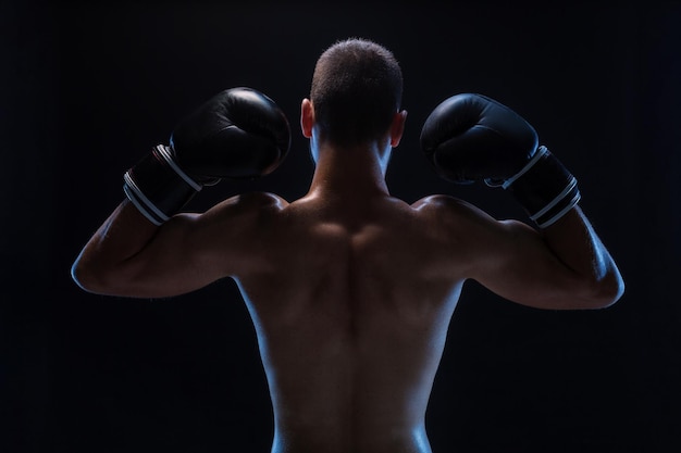 Rear view of strong young male boxer fitness male model wearing boxing gloves standing on black back...