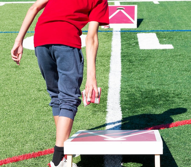 Rear view of a teenager throwing two bean hags while playing cornhole on a turf field