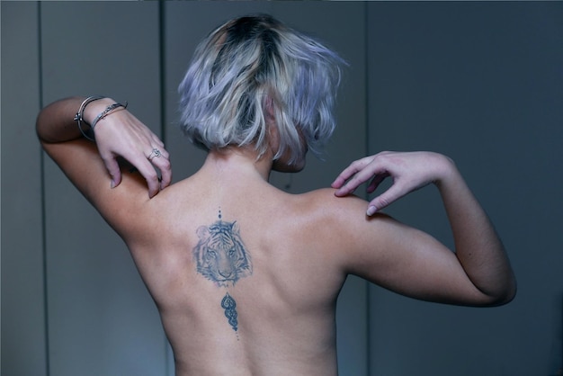 rear view of topless young woman with tattoo standing against wall