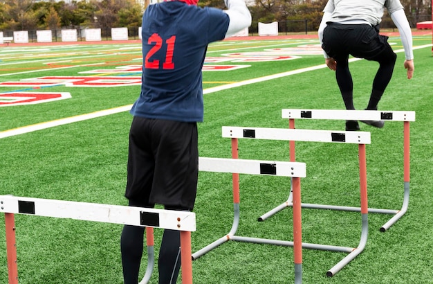 Rear view of two high school boys jumping over track hurdles on a green turf field