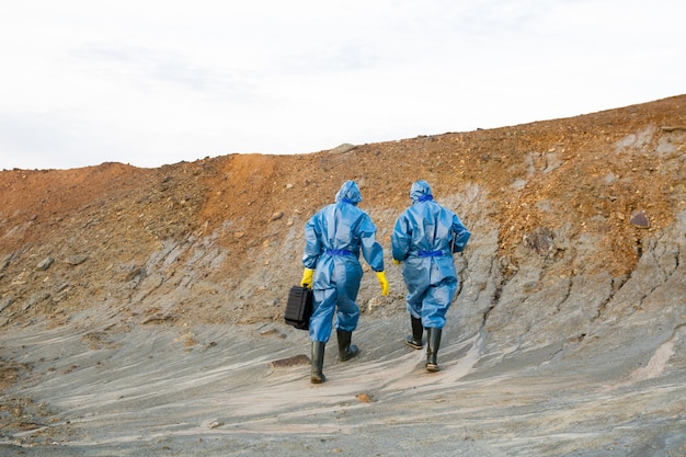 Rear view of two young contemporary ecologists in protective workwear ascending hill while investigating dangerous territory