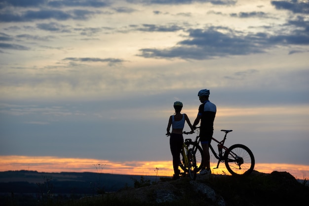 Rear view two young people with mountain bikes stand on top of cliff with beautiful scenery at sunset