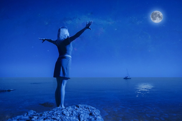 Фото Неопознанная молодая счастливая женщина, вид сзади, стоит на камне, поднимает руки вверх. глядя на большую луну и спокойную чистую морскую воду на фоне пейзажа и ясного ночного неба. концепция полной луны