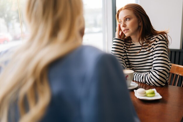 Задний вид неузнаваемой блондинки, разговаривающей с задумчивой подругой, проводящей время вместе во время питья кофе в уютном кафе, сидящей за столом у окна.