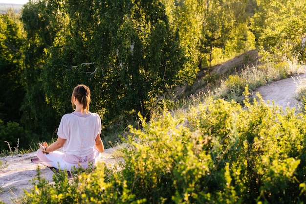 Rear view of unrecognizable young woman meditating in lotus position sitting on top of rock