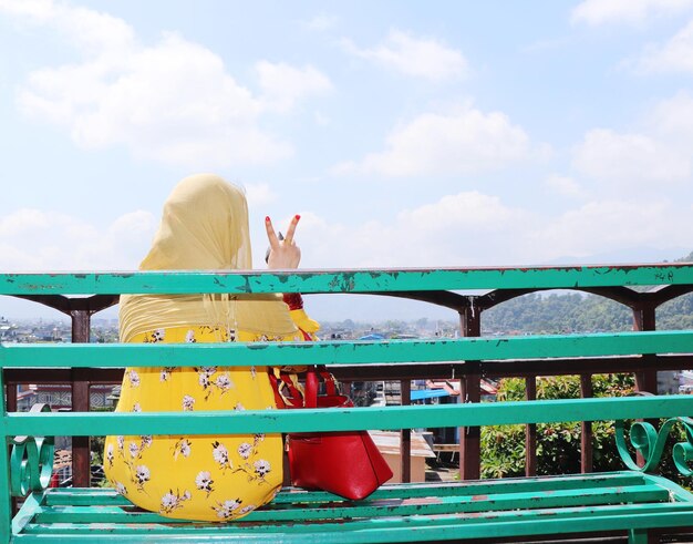 Rear view of woman gesturing while sitting on bench against sky