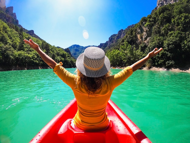 Rear view of woman in hat sitting on kayak with her arms raised
on a beautiful lake between mountain. woman enjoying kayaking
sailing on lake water surface during vacation