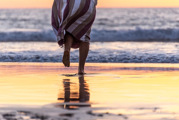 Rear view of woman legs walking on a beach to the ocean at\
sunset time.copy space