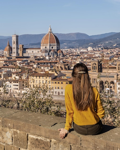 Foto vista posteriore di una donna che guarda il paesaggio cittadino mentre è seduta su un muro di sostegno