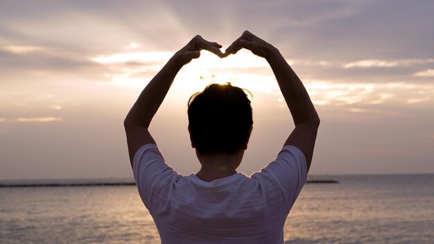 Foto vista posteriore di una donna che fa la forma di un cuore mentre si trova al mare contro il cielo