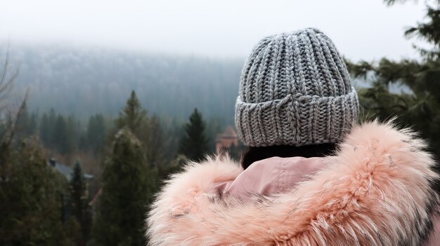 Vista posteriore di una turista di montagna donna in una giacca rosa con pelliccia e un cappello lavorato a maglia grigio su uno sfondo di montagne che guardano nel paesaggio autunnale della foresta e del cielo nuvoloso.