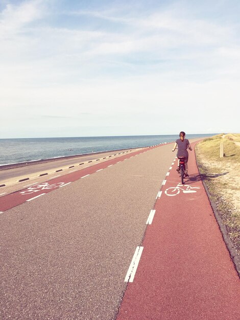 Photo rear view of woman riding bicycle on road by sea against sky during sunny day