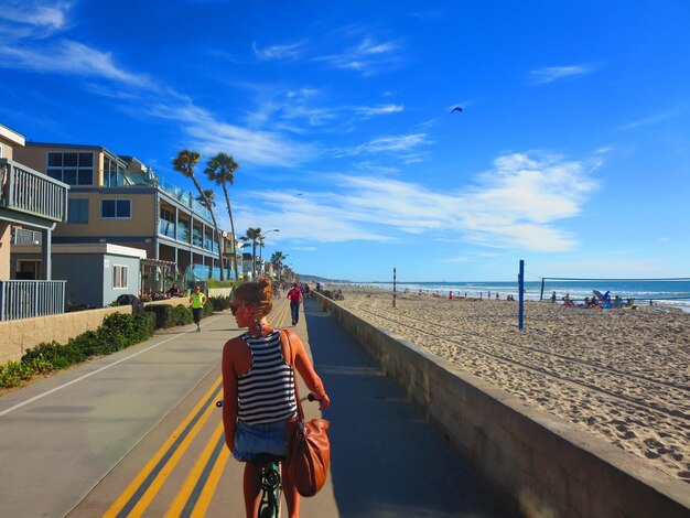 Задний вид женщины, едущей на велосипеде по тротуару на пляже на фоне голубого неба