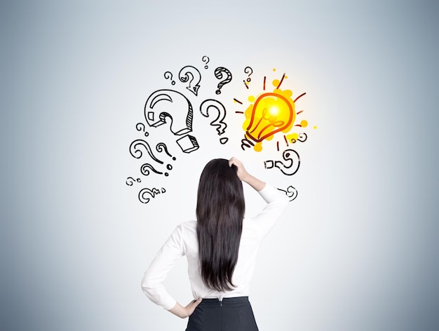 Rear view of a woman scratching her head and looking at a bright yellow light bulb sketch surrounded by question marks on a gray wall.