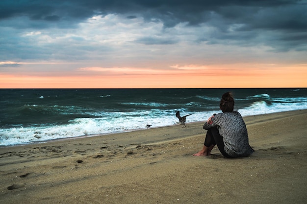 Foto vista posteriore di una donna seduta sulla spiaggia che guarda l'alba dell'oceano nuvole scure