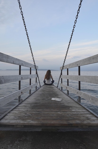 Foto vista posteriore di una donna seduta sul molo sopra il mare contro il cielo