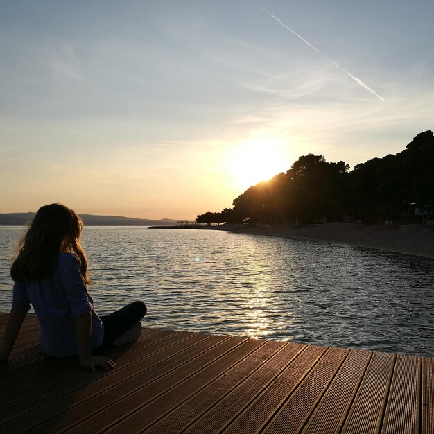 Foto vista posteriore di una donna seduta sul molo al lago durante il tramonto