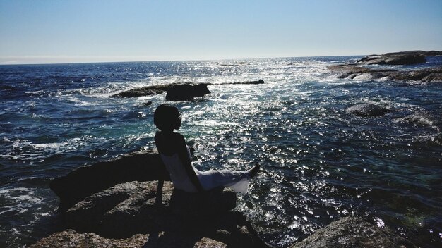 Foto vista posteriore di una donna seduta su una roccia in mare contro il cielo