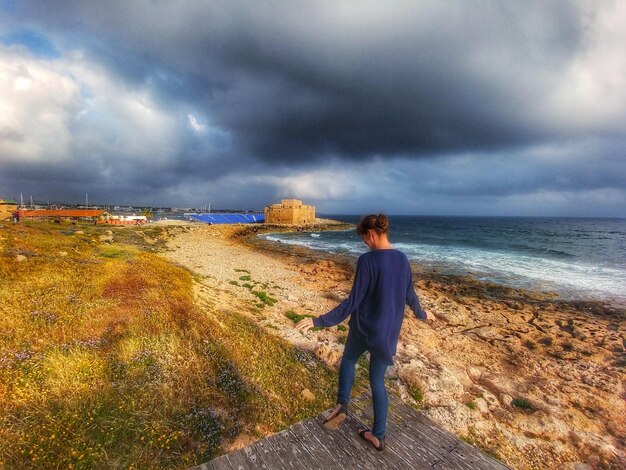 Rear view of woman standing on floorboard by sea against cloudy sky