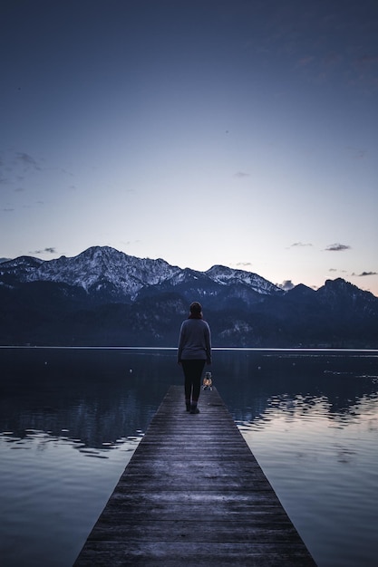 rear view of woman standing on pier by lake against sky during sunset