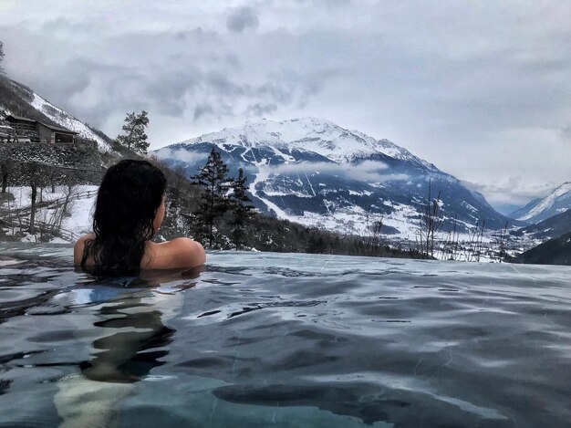 Foto vista posteriore di una donna che nuota in una piscina infinita contro montagne innevate