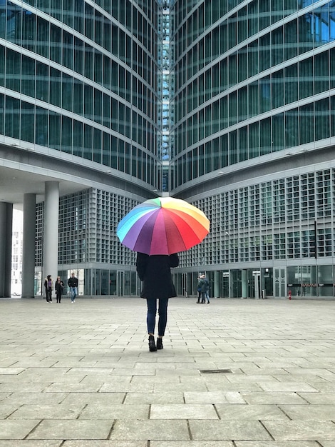 rear view of woman under umbrella standing on footpath by modern building in city