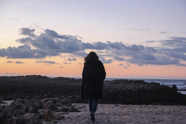 Rear view of woman walking at beach against sunset sky