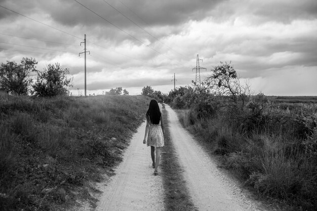 Photo rear view of woman walking on dirt road amidst plants against cloudy sky