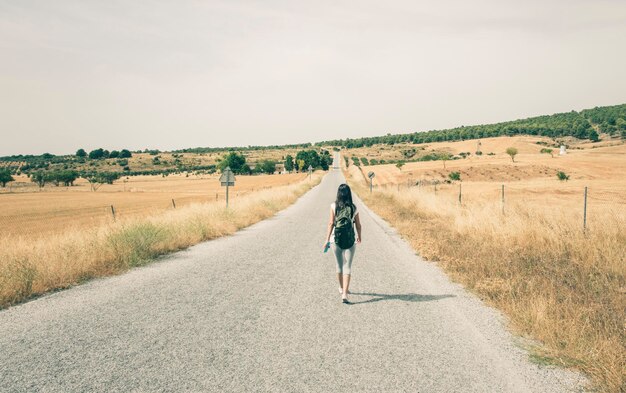 空の向こうのフィールドの真ん中に道路を歩く女性の後ろの景色