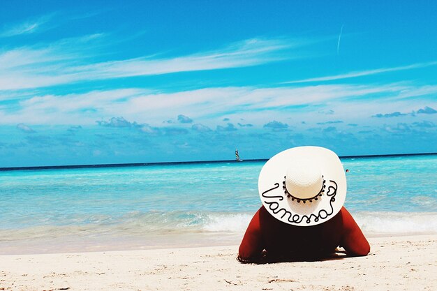 Rear view of woman wearing hat while resting at beach against sky during sunny day