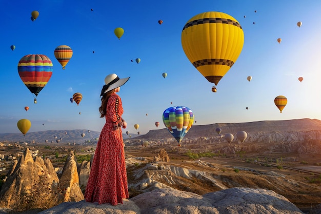 은 드레스와 모자를 입은 여성이 하늘을 향한 언덕에 서서 뜨거운 공기 풍선과 함께 뒷면