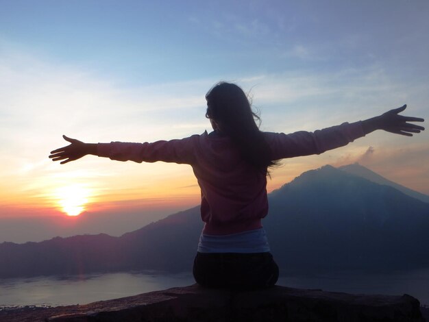 Photo rear view of woman with arms outstretched while sitting on rock against sky during sunset