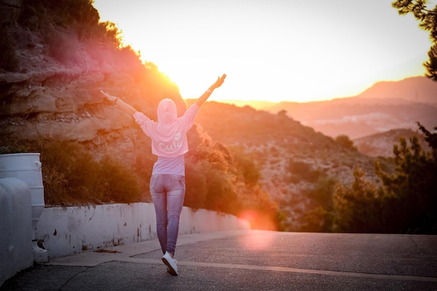 Foto vista posteriore di una donna con le braccia alzate in piedi sulla strada durante il tramonto