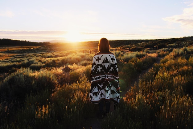 rear view of woman with blanket standing on field against sky during sunset