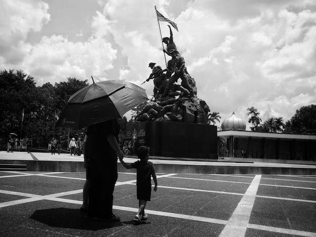 Foto vista posteriore di una donna con il figlio sotto un ombrello che cammina al memoriale di guerra