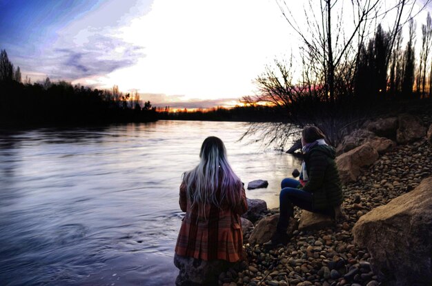 Foto vista posteriore di donne sedute vicino al lago contro il cielo