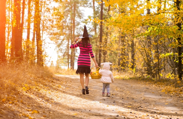 日差しの中で秋の林道を歩くハロウィーンパーティーのために服を着た子供を持つ女性の背面図