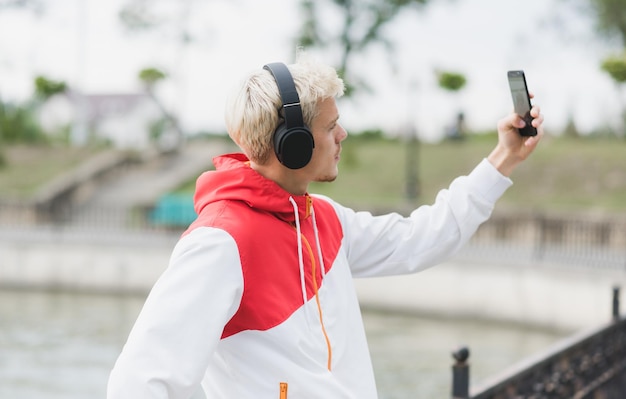 パーカーを着て、Bluetoothヘッドフォンで音楽を聴いている若い金髪のハンサムなヒップスターの男の背面図は、屋外でスマートフォンで自画像を撮る楽しみを持っています人々のライフスタイル技術の概念