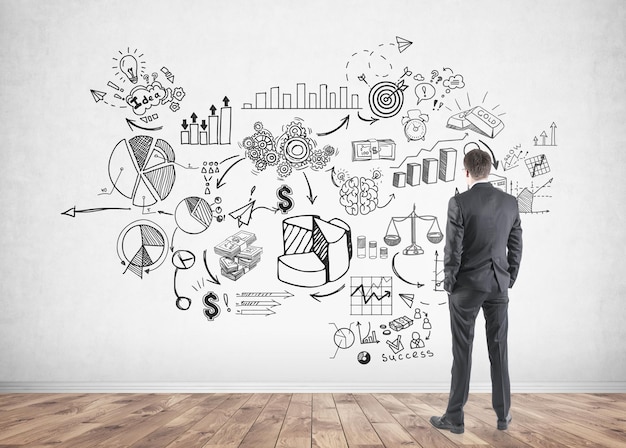 Rear view of a young businessman wearing glasses and a dark suit and looking at business plan sketch on a concrete wall.