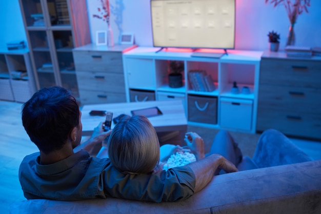 Вид сзади молодой пары, обнимающейся, сидя на диване, ест попкорн и смотрит фильм