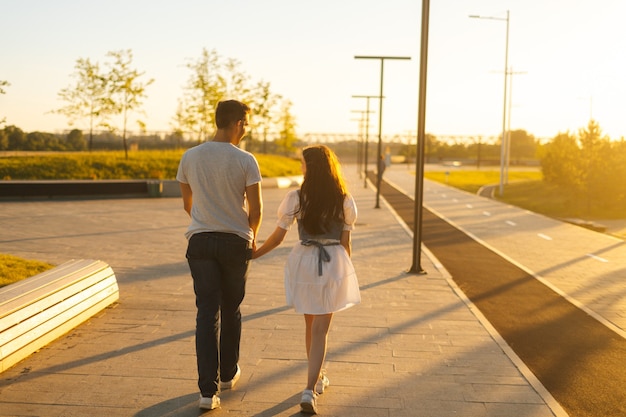 Rear view of young couple in love holding hands walking in city park in sunny summer day on background of bright sunlight. Happy relationships between boyfriend and girlfriend enjoying time together.