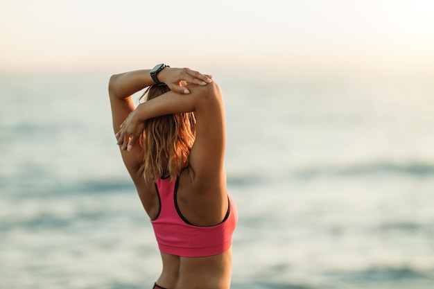 Вид сзади на молодую бегунью, выполняющую упражнения на растяжку во время тренировки возле морского пляжа.