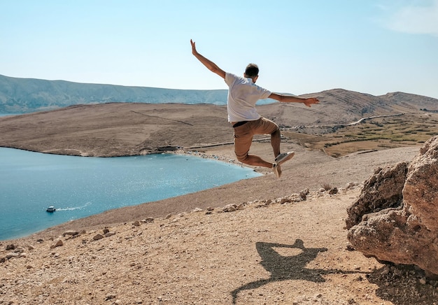 Вид сзади на молодого человека, прыгающего от радости на удивительном побережье с видом на залив с пляжем.