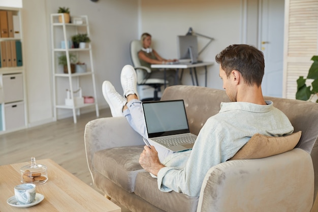 ソファに横になって、自宅の部屋でラップトップでオンラインで作業している若い男の背面図