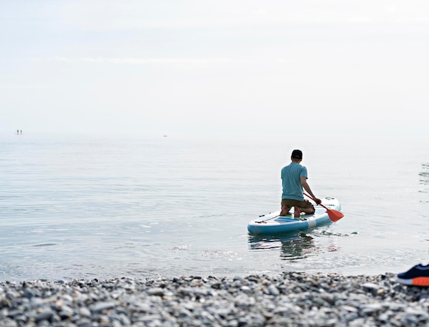Rear view of Young man sitting on blue stand up paddle board on sea on sunny summer day active lifestyle outdoor activity sup