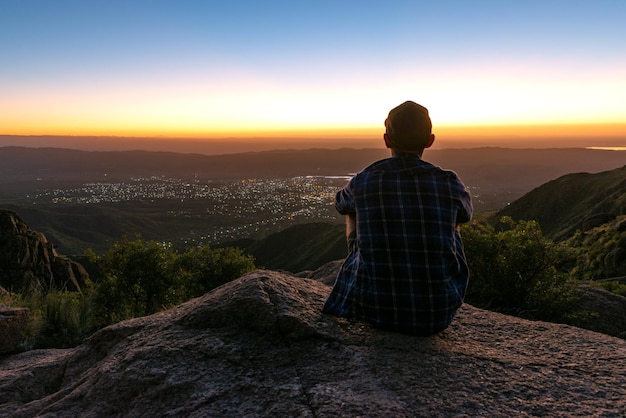 Вид сзади на молодого человека, сидящего на скале и наблюдающего закат за городскими огнями