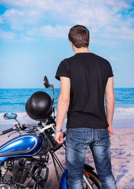 ビーチで古典的な青いオートバイの横に立って、水に面して若い男の背面図